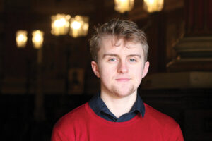 The photo shows a headshot of Exeter Cathedral Organ Scholar, Giles Longstaff, wearing a black shirt and red jumper. 