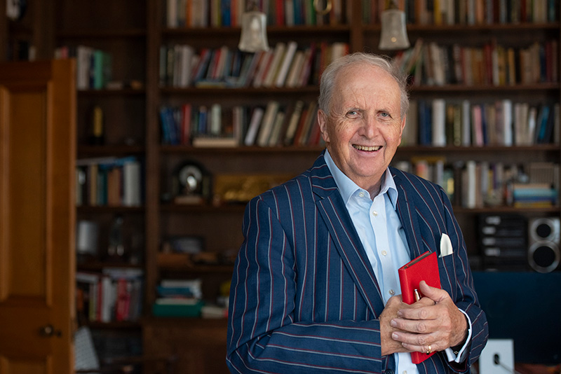Best-selling author, Alexander McCall Smith, is standing in front of a bookshelf with a smile on his face wearing a blue shirt, blue pinstripe blazer and holding a red book.