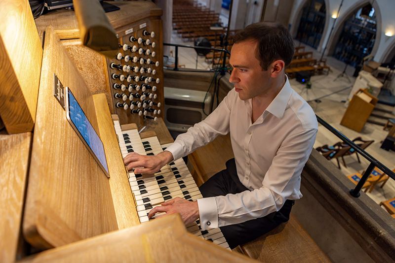 The photo shows Richard Moore, Sub-Organist at Christ Church Cathedral, playing an organ while reading the music from a tablet device. 