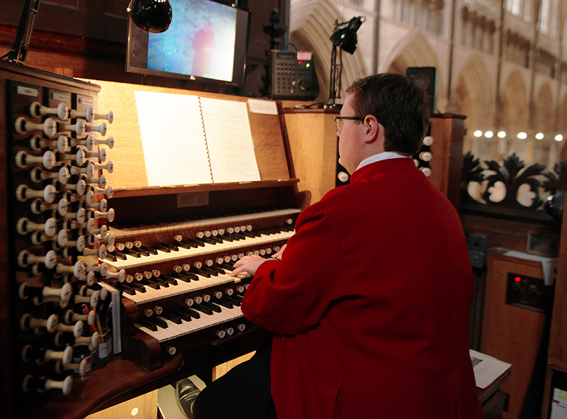 This photo shows Exeter Cathedral's Assistant Director of Music, Michael Stehpens-Jones, playing the Exeter Cathedral organ wearing a red cassock.