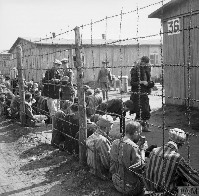 THE LIBERATION OF BERGEN-BELSEN CONCENTRATION CAMP, APRIL 1945. Prisoners sit by the wire fence dividing the various sections of the camp. They are eating their first meal after the liberation of the camp.