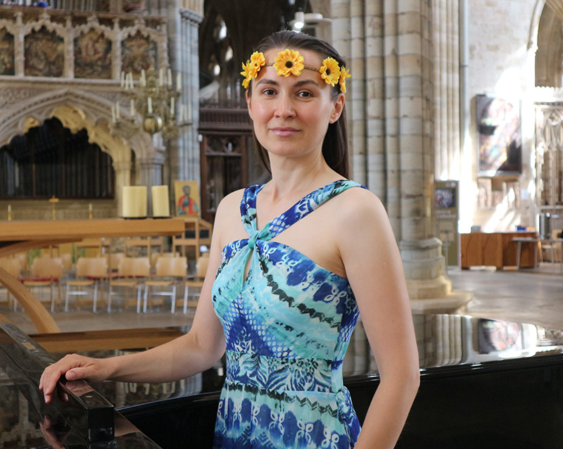 Ukrainian Mezzo-Soprano, Iryna Ilnytska standing in the nave of Exeter Cathedral in front of a grand piano wearing a blue summer dress and a sunflower headband.